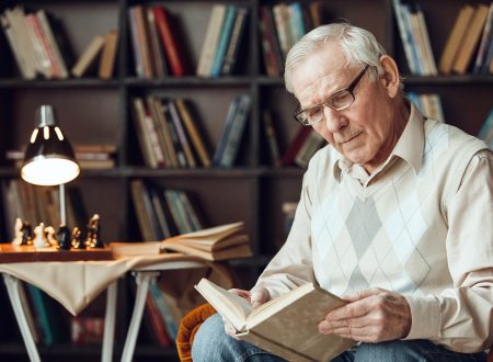 Senior man alone sitting on armchair at library wearing glasses reading book concentrated